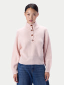 A model wearing a light pink ribbed knit mock neck sweater with four brown buttons, paired with blue jeans, against a light gray studio background.