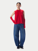 A model stands against a light gray studio background wearing a bright red sleeveless mock-neck knit top with matching red knit arm warmers, paired with loose blue jeans and black shoes.