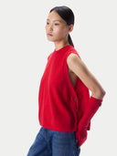 A model wearing a bright red sleeveless knit sweater vest with side buttons and a matching red knit sleeve cuff, paired with blue jeans, against a light gray studio background.