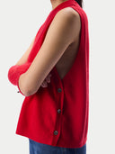 A close-up side view of a model wearing a bright red sleeveless knit top with a buttoned front and a large open back, paired with blue jeans against a light gray studio background.