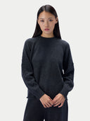 A model wearing a dark charcoal gray knit crewneck sweater and matching dark pants, standing against a light gray studio background.