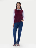 A model stands against a light gray studio background wearing a sleeveless burgundy knit sweater vest over a white collared shirt, blue jeans, and dark heeled sandals.
