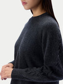 A close-up side view of a woman wearing a dark charcoal gray knit sweater with textured cable detailing on the sleeve, set against a plain light gray background.