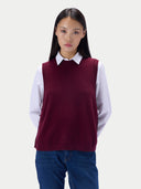 A model wearing a sleeveless burgundy knit sweater vest over a white collared shirt and blue jeans, photographed against a light gray studio background.