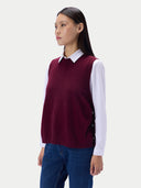 A woman wearing a sleeveless burgundy knit sweater vest over a white collared shirt and blue jeans, photographed against a light gray studio background.