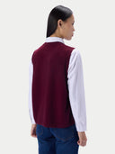 A woman shown from the back wearing a sleeveless burgundy knit vest over a white collared shirt and blue jeans, against a plain light gray studio background.