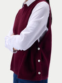 A person wearing a sleeveless burgundy knit sweater vest over a white collared shirt, paired with blue jeans, shown from the side against a light gray background.