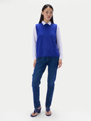 A model stands against a light gray studio background wearing a royal blue knit sweater vest over a white collared shirt, paired with dark blue jeans and dark blue heeled sandals.