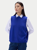 A woman wearing a royal blue knit sweater vest over a white collared shirt and dark blue jeans, photographed against a light gray studio background.