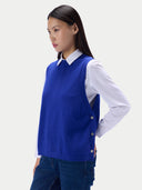 A woman wearing a royal blue sleeveless knit sweater vest over a white collared shirt and dark blue jeans, photographed against a light gray studio background.