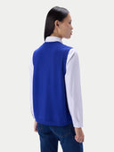 A woman shown from the back wearing a royal blue knit sweater vest over a white collared shirt and dark blue jeans, against a light gray studio background.