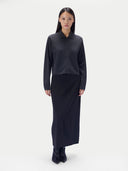 A model stands against a light gray studio background wearing a dark charcoal gray long-sleeve collared knit top and a matching dark navy or black long skirt, paired with black ankle boots.