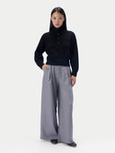 A model stands against a light gray studio background wearing a black long-sleeve mock-neck knit top with a button placket and high-waisted, wide-leg gray trousers.