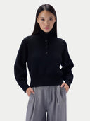 A woman modeling a black ribbed mock-neck knit sweater with three front buttons, paired with gray trousers, against a light gray studio background.