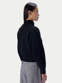 A woman shown from the back and side wearing a dark navy ribbed knit mock-neck sweater and light gray trousers, photographed against a clean light gray studio background.