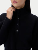 Close-up of a model wearing a black ribbed knit mock-neck sweater with a buttoned placket and dark buttons, against a light gray background.