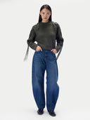 A model stands against a light gray studio background wearing a dark olive green ribbed knit sweater with fringe details on the sleeves and wide-leg blue jeans.