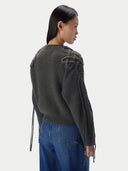 A woman shown from the back wearing a dark olive green ribbed knit sweater with knotted decorative details on the shoulders and long tie strings hanging from the sleeves, paired with blue jeans against a light gray studio background.