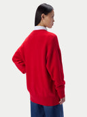 A woman wearing a bright red knit cardigan over a white collared shirt and blue jeans, photographed against a clean light gray studio background.
