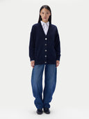 A model stands against a light gray studio background wearing a dark navy cardigan over a white collared shirt, paired with loose blue jeans and black shoes.