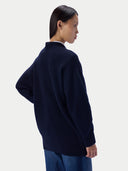 A model shown from the back wearing an oversized navy blue knit sweater over a white collared shirt and blue jeans, set against a plain light gray studio background.