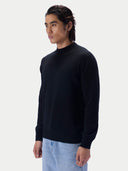 A man wearing a black mock neck knit sweater and light blue jeans, standing against a plain light gray studio background.