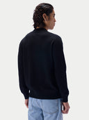 A man shown from the back wearing a black knit sweater and light blue jeans against a plain light gray studio background.