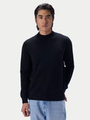 A man wearing a black long-sleeve mock neck knit sweater and light blue jeans, standing against a light gray studio background.