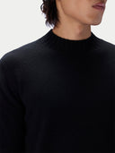 Close-up of a person wearing a black ribbed mock neck sweater against a light gray background.