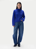 A model stands against a light gray studio background wearing a bright royal blue mock-neck knit sweater with a braided front detail and wide-leg blue jeans.
