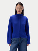 A model wearing a royal blue mock-neck knit sweater with a braided front detail, paired with blue jeans, against a light gray studio background.