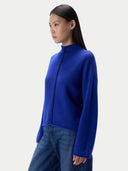 A woman wearing a bright royal blue mock-neck knit sweater with a braided front detail and blue jeans, photographed against a clean light gray studio background.