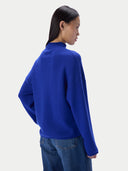 A woman shown from the back wearing a bright royal blue mock-neck knit sweater and blue jeans against a plain light gray studio background.