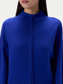 A close-up of a model wearing a vibrant royal blue knit sweater with a mock neck and a central braided cable detail running down the front.