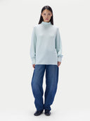 A model stands against a plain light gray studio background wearing a turtleneck sweater, loose dark blue jeans, and black shoes.