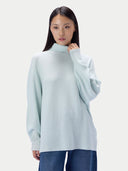 A woman modeling an oversized turtleneck sweater with wide, draped sleeves, paired with blue jeans, against a solid off-white background.