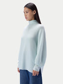 A woman wearing a morning green turtleneck sweater and blue jeans, shown in profile against a light gray studio background.