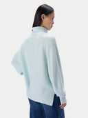 A model shown from the back wearing a pale mint green mock-neck knit sweater and dark blue jeans against a light gray studio background.