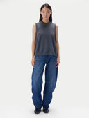 A model stands against a light gray studio background wearing a dark gray sleeveless mock-neck knit top and loose-fit blue jeans.