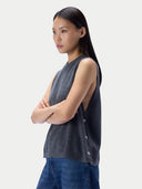 A woman wearing a dark gray sleeveless knit top with side buttons and blue jeans, photographed against a light gray studio background.