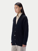 A woman wearing a dark navy blue knit cardigan over a cream collared shirt and light beige trousers, photographed against a solid light gray background.