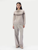 A model stands against a plain light gray studio background wearing a light beige knit sweater with a dark brown geometric Fair Isle pattern across the chest and shoulders, paired with matching light beige wide-leg trousers.
