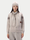 A model wearing a light beige knit sweater, matching knit pants, a knit beanie, and a matching knit scarf with dark geometric patterns and a small brown brand patch.