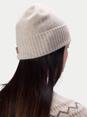 A woman shown from the back wearing a light beige knit beanie with a small brown brand tag on the side. The beanie has a ribbed cuff and is photographed against a clean, light gray background.