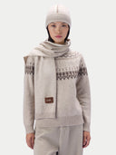 A model wearing a light beige knit sweater with a dark brown Fair Isle pattern on the yoke, a matching knit scarf, a knit beanie, and light gray trousers. A small brown brand patch on the scarf reads GOBI.