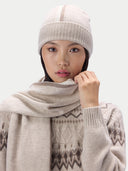 A woman wearing a light beige knit beanie, a matching soft scarf, and a patterned knit sweater against a plain light gray background.