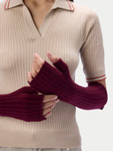 A close-up of a person wearing ribbed fingerless knit arm warmers in deep burgundy over a beige ribbed knit polo sweater with thin red stripe accents on the collar and cuffs.