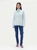 A woman wearing a light mint green loose-fit turtleneck sweater, dark blue jeans and navy sandals, standing against a light gray studio background.