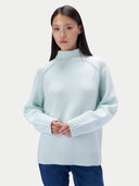 A woman wearing a light mint green turtleneck knit sweater with raglan sleeves and dark blue jeans, standing with her hands clasped against a light gray studio background.