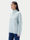 A woman shown in side profile wearing a light mint green turtleneck knit sweater with ribbed cuffs and hem and dark blue jeans, against a light gray studio background.